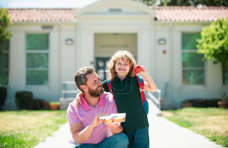 Photo for Parent and pupil of primary school go hand in hand. Schoolboy and parent in shirt holding lunch box. Teacher in t-shirt and cute schoolboy with backpack near school park - Royalty Free Image
