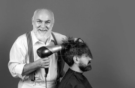 Téléchargez les photos : Homme barbu obtenir coiffure par coiffeur avec sèche-cheveux au salon de coiffure. Coiffeur heureux tenant un sèche-cheveux - en image libre de droit