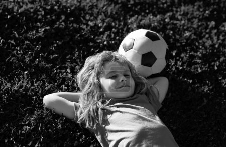 Téléchargez les photos : Garçon de football rêvant, allongé sur l'herbe. Jeune footballeur. Du sport pour les enfants. Rêver enfants se détendre en plein air - en image libre de droit