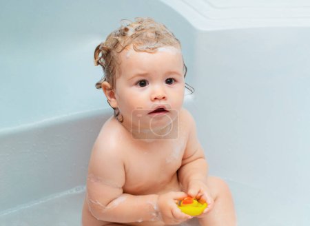 Téléchargez les photos : Bains de bébé drôles dans la baignoire avec de l'eau et de la mousse. Hygiène des enfants. Petit enfant dans une baignoire. Enfant souriant dans la salle de bain avec canard jouet - en image libre de droit