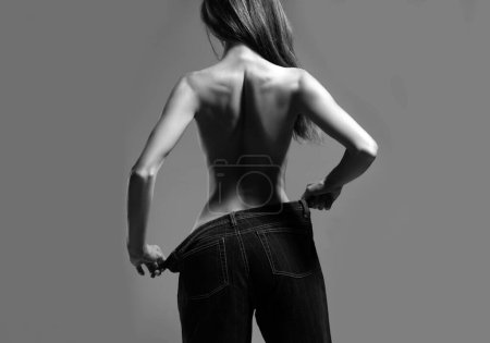 Téléchargez les photos : Slim girl have points at slim waist in big trousers, successful weight loss, isolated on gray background. Successful weight loss, woman with too large jeans after a diet. Skinny waist, back view - en image libre de droit