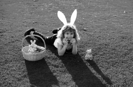 Foto de Niño con huevos de Pascua y orejas de conejito poniendo en la hierba. Feliz Pascua - Imagen libre de derechos
