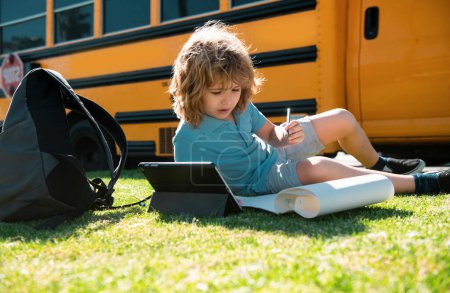Téléchargez les photos : Écolier écrivant en plein air dans le parc scolaire et faisant des devoirs sur le bus scolaire - en image libre de droit