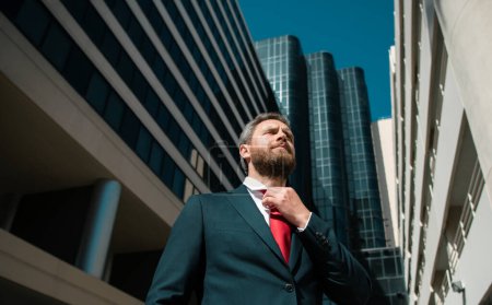 Téléchargez les photos : Homme d'affaires en costume bleu attachant la cravate en plein air. Homme d'affaires bien habillé sur l'immeuble de bureaux de la ville - en image libre de droit