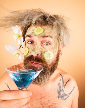 Téléchargez les photos : Funny man in hawaii. Male model with clay mask and cucumber slices on face drink cocktail. Man having cosmetic moisturizing mask hawaiian flowers - en image libre de droit