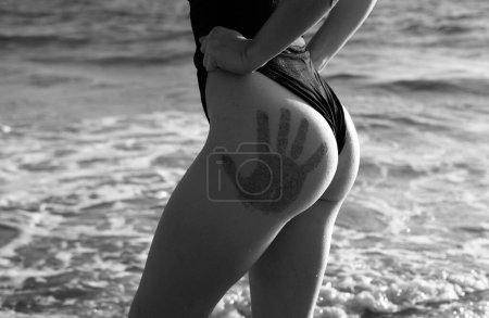 Foto de Chica con botín sexy con huellas de manos en bikini negro descansando en la playa desierta. Hermosa modelo en traje de baño en arena tropical cerca del mar - Imagen libre de derechos