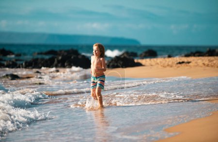 Téléchargez les photos : Petit garçon courant sur le rivage de la plage éclaboussant l'eau dans la mer bleue. Enfant marchant sur la plage d'été - en image libre de droit
