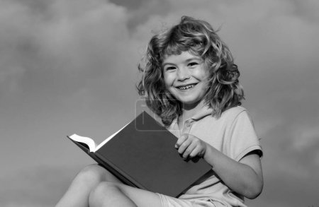 Foto de Lindo libro de lectura de niños encantadores en el fondo azul del cielo exterior. Niño leer libro en el parque - Imagen libre de derechos