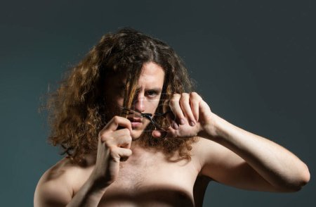 Foto de Joven hombre sexy, retrato de tipo con tijeras de peluquero corte pelo largo rizado para peluquería. Peluquería moderna, afeitado. Hombre guapo con peinado moderno en negro - Imagen libre de derechos