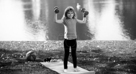 Téléchargez les photos : Sport pour enfants. Enfant sain garçon avec haltère exercice en plein air. Petit enfant pratique haltères exercices dans le parc - en image libre de droit