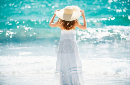 Téléchargez les photos : Vue arrière belle fille avec chapeau de paille profitant du bain de soleil à la plage. Jeune femme bronzée profitant de la brise au bord de la mer. Femme insouciante souriant avec océan de mer en arrière-plan - en image libre de droit