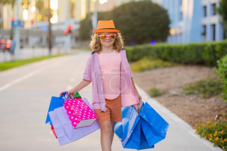 Photo for Cute little boy in summer fashion clothes goes shopping. Happy child with shopping packages in hands. Shopper child with shopping bag outdoor. Cute kid walking on street carrying shopping bags - Royalty Free Image