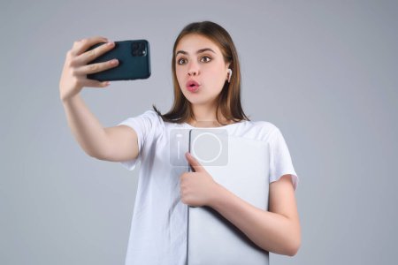 Photo for Young woman Student in white t-shirt holding phone and laptop, isolated over gray background. Student making video call. Girl Student with smart phone in studio. Portrait of student having video-call - Royalty Free Image