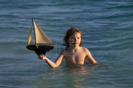 Téléchargez les photos : Petit garçon profitant de vacances d'été au bord de la mer. Un petit garçon mignon jouant avec un bateau jouet sur la plage par une chaude journée ensoleillée d'été. Vacances en mer. Enfant jouant avec un voilier jouet. Journée des enfants - en image libre de droit