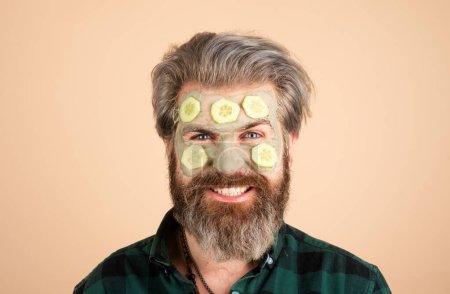Téléchargez les photos : Homme avec masque facial amusant avec concombre. Spa homme appliquant masque d'argile faciale. Spa à domicile. Hommes tenant des morceaux de concombre sur leur visage, isolés - en image libre de droit