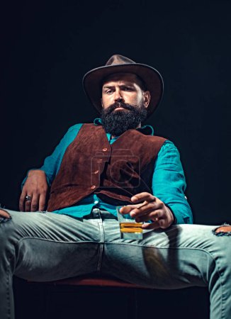 Téléchargez les photos : Homme d'affaires barbu en costume élégant avec un verre de whisky. Concept de style mode. L'homme barbu joyeux boit du whisky cher - en image libre de droit