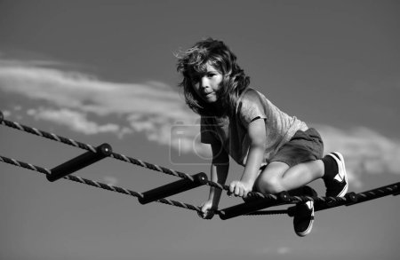 Photo for Kid climbing the net. Cute boy climbs up the ladder on the playground. Child climbs up the ladder against the blue sky - Royalty Free Image