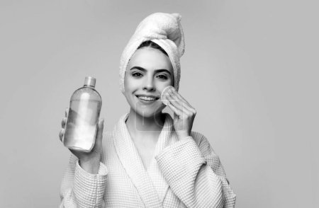 Foto de Chica con agua micelar. Hermosa mujer con toalla en la cabeza, agua micelar y almohadilla de algodón aislado en gris - Imagen libre de derechos