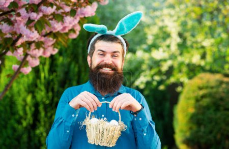 Foto de Retrato del hombre de Pascua cazando huevos. El hombre con orejas de conejo sostiene la cesta con huevo de Pascua. Caza de huevos. Vacaciones de primavera - Imagen libre de derechos