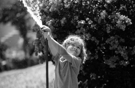 Foto de Niño jugando con manguera de jardín en el patio trasero. Divertido niño emocionado divertirse con spray de agua en el fondo de la naturaleza patio. Actividades al aire libre para niños de verano - Imagen libre de derechos