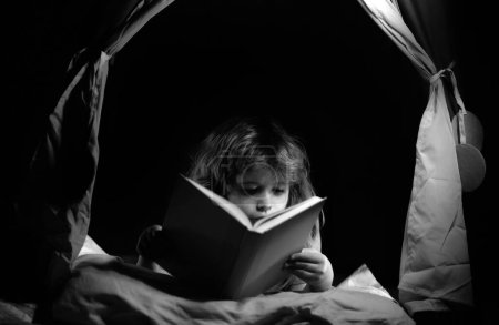 Téléchargez les photos : Des enfants dans une tente pour enfants lisant un livre dans le noir. Enfant lisant un livre allongé sur le lit. Rêver enfant lire des histoires au coucher, conte de fées ou conte de fées - en image libre de droit