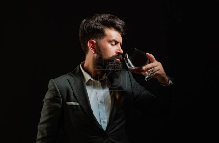 Téléchargez les photos : Élégant homme barbu élégant Barman tient verre de whisky. Sommelier goûte boisson alcoolisée coûteuse - en image libre de droit