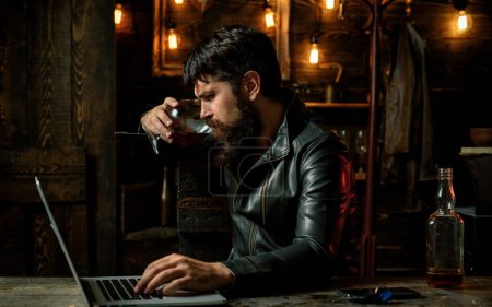 Téléchargez les photos : Élégant homme en veste de cuir buvant un whisky dans un bar. Un hipster brutal qui boit de l'alcool. élégant beau barbu homme appréciant whisky et faire ses affaires à l'ordinateur portable - en image libre de droit