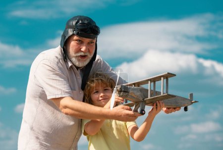 Téléchargez les photos : Vieux grand-père et jeune petit-fils enfant jouant avec avion en bois sur fond de ciel d'été. Enfant garçon avec des rêves de voler ou de voyager - en image libre de droit