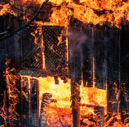 Foto de Un incendio destruye una casa quemada carbonizada. Quemado en la casa de la llama por la noche. Fuego por todas partes y humo en una zona residencial por la noche. Fuego peligroso. Toda la casa y el patio en llamas - Imagen libre de derechos