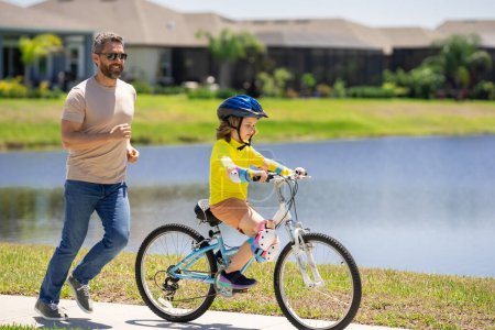Téléchargez les photos : Père et fils à vélo sur un parc. Enfant en casque de sécurité avec son père à vélo le jour de l'été. Père enseignant fils à vélo. Enfant fils en casque de vélo et père à vélo. Fête des pères - en image libre de droit