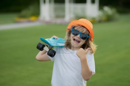 Téléchargez les photos : Joyeux garçon riant et tenant le skateboard en plein air. Portrait d'été pour enfants. Drôle excité enfant avec le pouce levé. Enfant excité - en image libre de droit