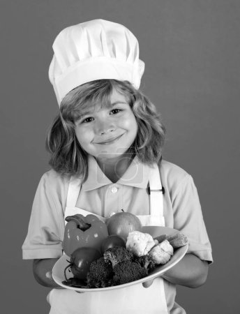 Téléchargez les photos : Chef enfant garçon tenir assiette avec des légumes faisant des légumes frais pour une alimentation saine. Portrait de petit enfant en forme de cuisinier isolé sur fond bleu. Un petit chef. Processus de cuisson - en image libre de droit