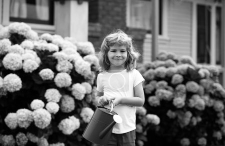 Foto de Lindo niño regando flores con regadera en el jardín. Niño vestido en verano ligero cierra y colorido camiseta, sonriendo y divirtiéndose. Actividades con niños al aire libre - Imagen libre de derechos