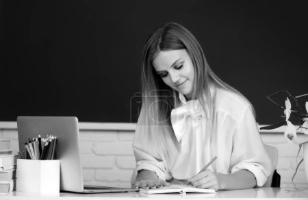 Photo for Focused student, young woman, writing on notebook learning education course, sit at work desk in classroom, elearning concept - Royalty Free Image