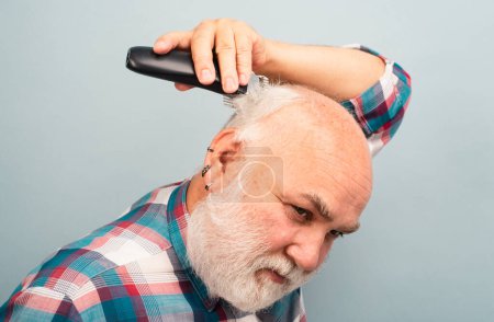 Téléchargez les photos : Un barbu se coupe les cheveux avec une tondeuse. Traitements capillaires. Coupes de cheveux d'homme gris. Tondeuse homme chauve, Calvitie mature et concept de perte de cheveux - en image libre de droit