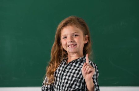 Photo for Portrait of school girl nerd pupil with surprising expression pointing with finger against blackboard. Back to school, first day at school - Royalty Free Image
