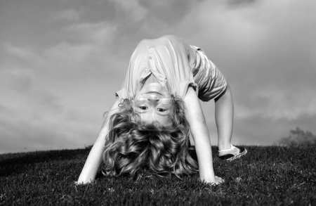 Foto de Niño feliz niña de pie boca abajo sobre su cabeza en la hierba en el día de verano. Divertido lindo niño haciendo ejercicio divertido - Imagen libre de derechos