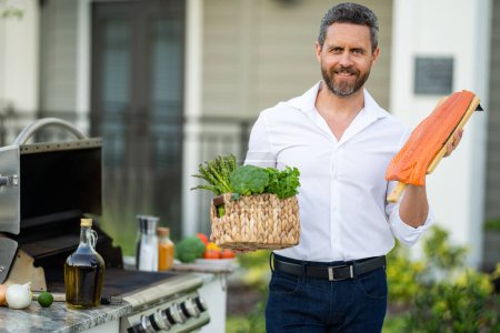 Photo for Man at a barbecue grill. Male cook preparing barbecue salmon fillet outdoors. Bbq fish fillet, grill for picnic. Roasted salmon. Attractive man preparing barbeque salmon fillet in the house yard - Royalty Free Image