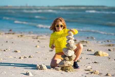 Téléchargez les photos : Méditation estivale pour enfants. Enfant garçon avec pyramide de galets en pierre équilibrée sur la plage. Jeu d'enfants avec des pierres zen sur la plage de la mer, méditation, spa, harmonie, calme et équilibre concept pour les enfants - en image libre de droit