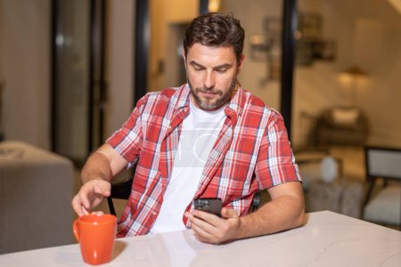 Photo for Portrait of man looking at smart phone at living room. Man is talking hold cup of coffe chatting on the smartphone. Thoughtful man using mobile phone at home. Middle-aged man using phone - Royalty Free Image