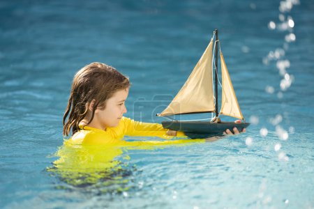 Téléchargez les photos : Petit garçon blond mis bateau jouet dans les vagues de la mer à la plage pendant les vacances d'été. Vacances d'enfance et d'été en famille. Rêve en voyage. Visite guidée en voilier. Concept de rêve enfants - en image libre de droit