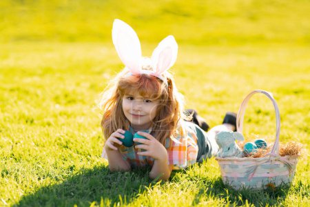 Téléchargez les photos : Enfant pondant sur l'herbe dans les oeufs de Pâques parc esprit. Lapin de Pâques enfants couchés sur l'herbe. Enfants garçon dans les oreilles de lapin chasse oeufs de Pâques dans le parc en plein air - en image libre de droit