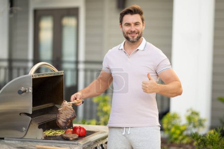 Photo for Man at a barbecue grill. Male cook preparing barbecue outdoors. Bbq meat, grill for picnic. Roasted beef. Cook preparing barbeque in the house yard. Barbecue and grill. Cook using barbecue tongs - Royalty Free Image