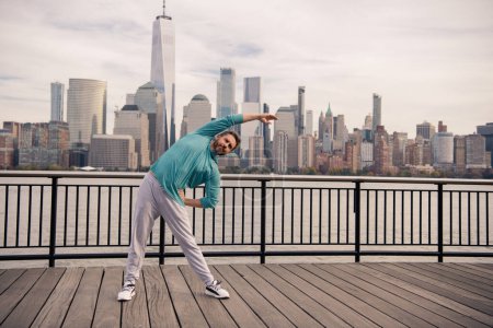 Foto de Hombre en entrenamiento de ropa deportiva cerca de Manhattan. Un estilo de vida saludable. Hombre mayor activo es ejercicio de fitness al aire libre. Fitness después de la jubilación. Maduro deportista retirado haciendo ejercicios de estiramiento - Imagen libre de derechos