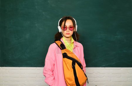 Foto de Joven adolescente colegiala con la mochila de la escuela y auriculares. Adolescente estudiante en pizarra - Imagen libre de derechos