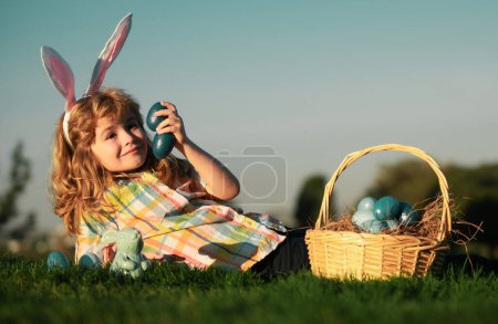 Téléchargez les photos : Enfant garçon avec des œufs de Pâques dans le panier en plein air. Un gamin étendu sur l'herbe dans le parc. Chasse aux œufs de Pâques. Les enfants de Fynny font face. Mignon garçon lapin enfant avec des oreilles de lapin sur fond de ciel avec espace de copie - en image libre de droit
