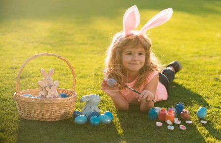 Foto de Niño con huevos de Pascua y orejas de conejito poniendo en la hierba. Chico divertido, conejito de Pascua niños - Imagen libre de derechos