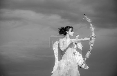 Téléchargez les photos : Fille en robe d'anges blancs, Cupidon avec des ailes visant avec arc et flèche sur un fond de ciel. Concept de Saint Valentin. ange adolescent dans ciel - en image libre de droit