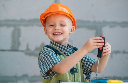 Foto de Kid boy twists bolt with screwdriver. Child repairman with repair tool. Child in helmet and boilersuit on construction site. Little worker engineer - Imagen libre de derechos