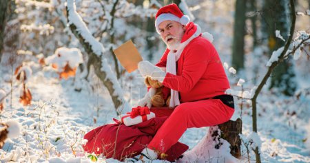 Téléchargez les photos : Père Noël lire la liste de souhaits de Noël sur le fond extérieur d'hiver. Le Père Noël porte un cadeau de Noël. Père Noël sur fond de Noël. Père Noël âgé avec des cadeaux et des jouets - en image libre de droit